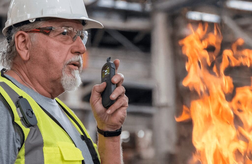 Older industrial worker wearing a hard hat and safety vest uses a Relay device to communicate near an active fire, highlighting emergency communication in hazardous environments.