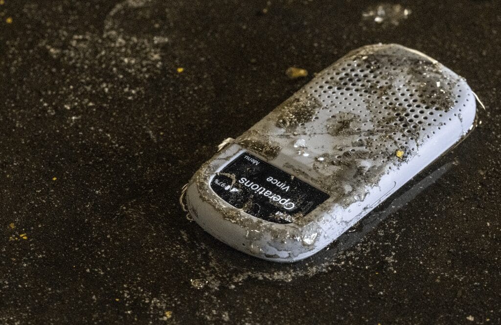 A Relay communication device covered in dirt and water lies on a wet, gritty industrial floor, displaying a message from “Operations – Vince” on the screen.