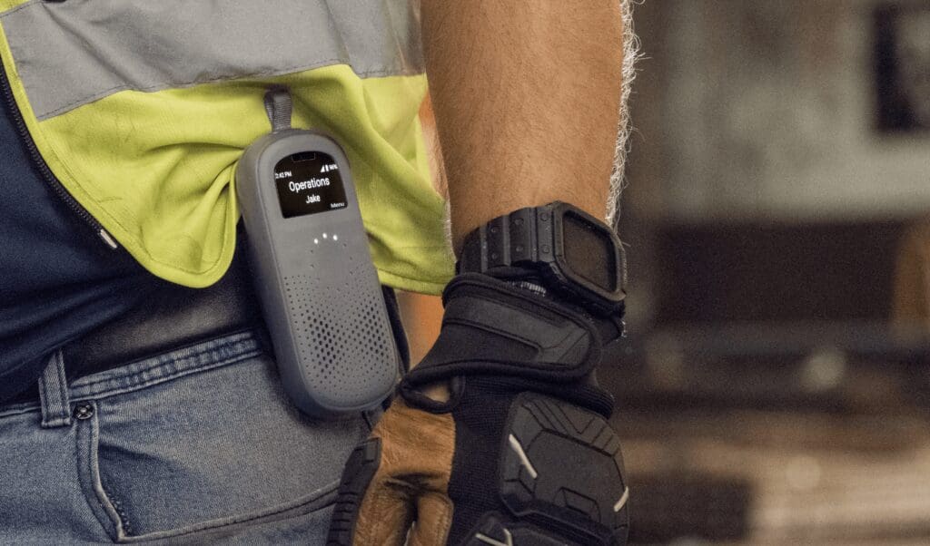 Close-up of a worker wearing a safety vest and gloves with a Relay communication device clipped to their belt, displaying an active channel labeled "Operations – Jake."
