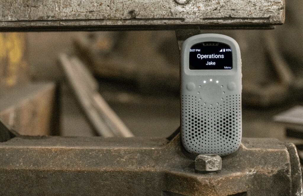 A Relay communication device rests on a piece of industrial equipment in a workshop setting, displaying a message from “Operations – Jake” on its screen.