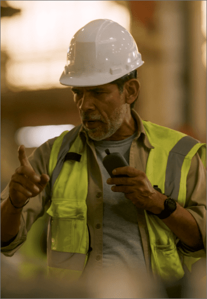 Male supervisor in a high-visibility vest and hard hat speaking into a Relay device on the manufacturing floor.