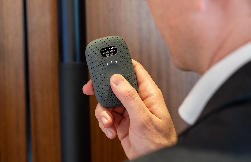 Person in a suit holding a gray RelayM device, pressing the button to send a message, with the screen displaying “Operations – Richard.”