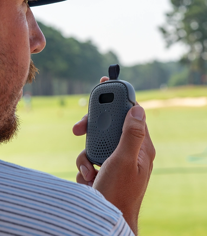 Person holding a Relay device outdoors, speaking into it, with a golf course visible in the blurred background.