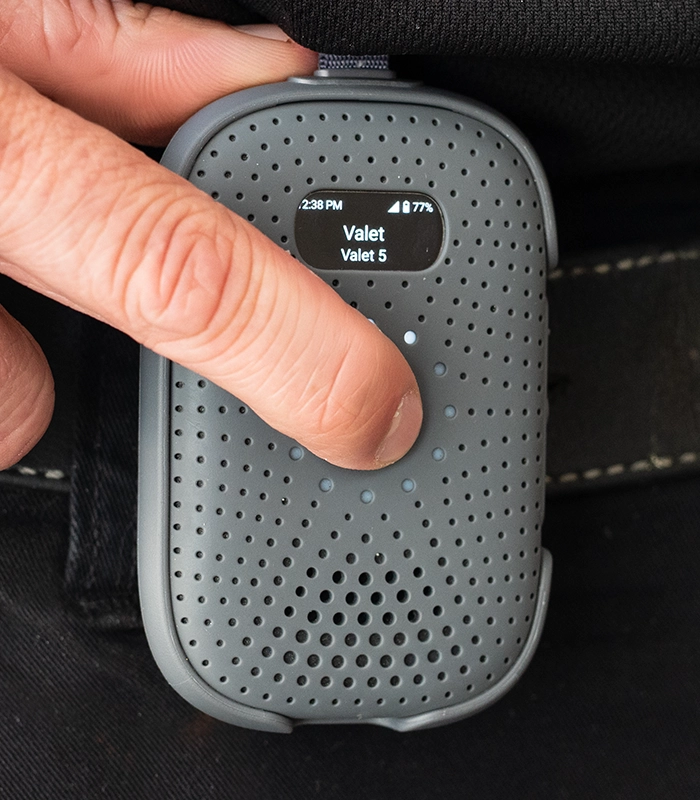 Close-up of a person pressing the talk button on a Relay M device clipped to their waistband, with the display showing “Valet” and battery level.