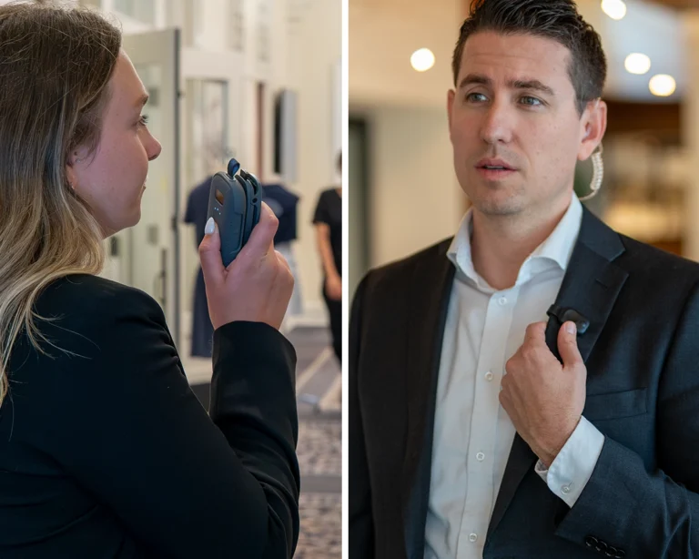 Two security professionals using Relay communication devices — one woman speaking into a handheld RelayX and one man using an earpiece-connected Relay device — in a hotel or event venue setting for real-time coordination.